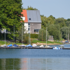Gästehaus Schnapp´s Hof am Möhnesee Haus am See mit Booten am Steg und bewaldeter Uferpromenade.