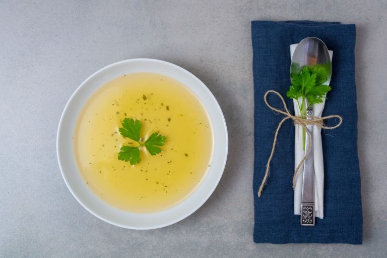 Fastenguide Klare Suppe mit Kräutern auf einem grauen Untergrund, Besteck daneben.